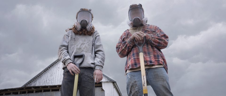 Two figures in protective masks and hoods walk a farm road under a stormy sky.