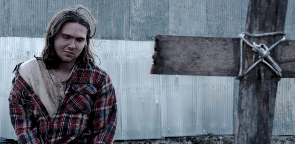 Jude, played by Zach Hurley, kneeling beside a wooden cross marker.