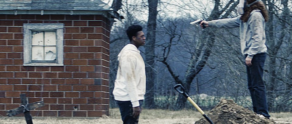 A masked figure raises a handgun near a grave marker and a brick shed.