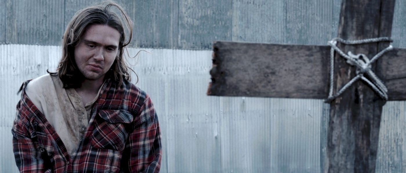 Jude (Zach Hurley) sits beside a wooden cross marker.