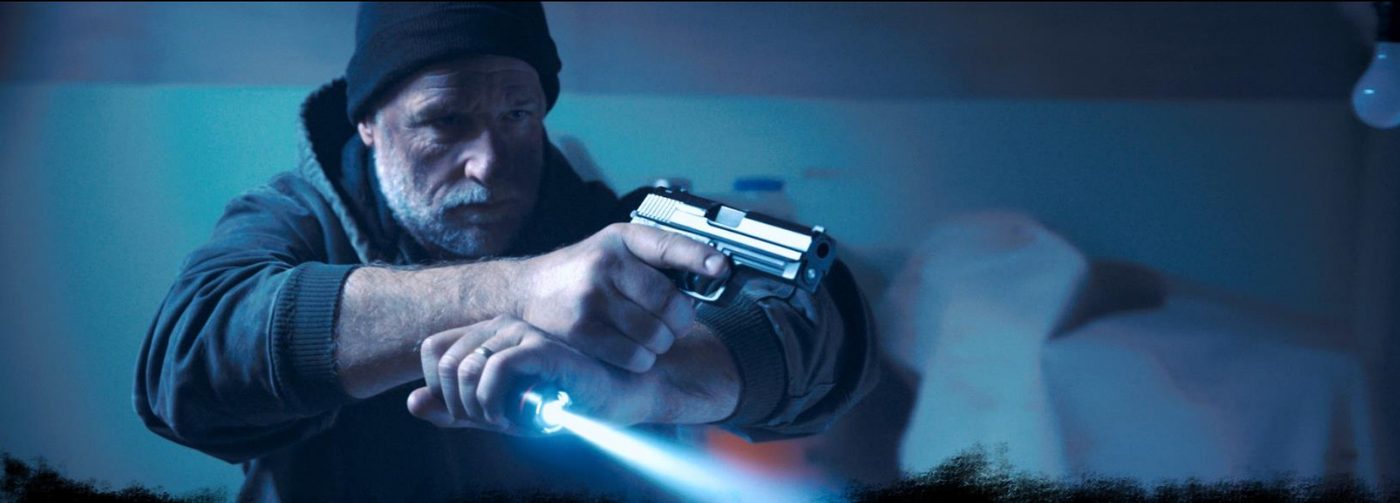 Mac, played by Daniel J. Lennox, aims a pistol and flashlight into the dark.