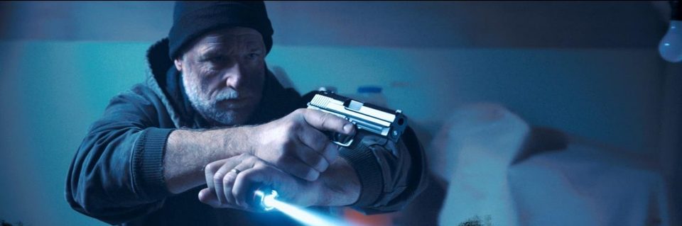 Mac, played by Daniel J. Lennox, aims a pistol and flashlight into the dark.